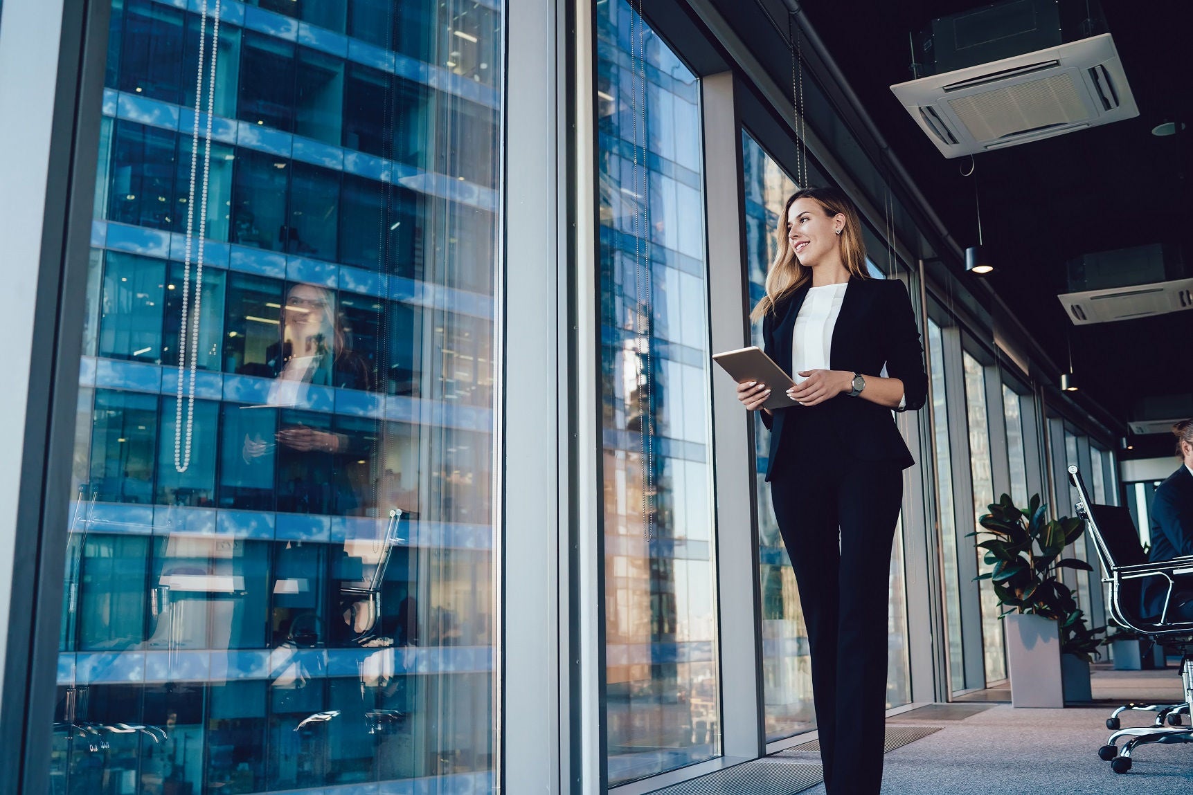 Caucasian female entrepreneur dressed in formal wear going near panoramic window in office, confident business woman with modern digital tablet looking away during working day in enterprise