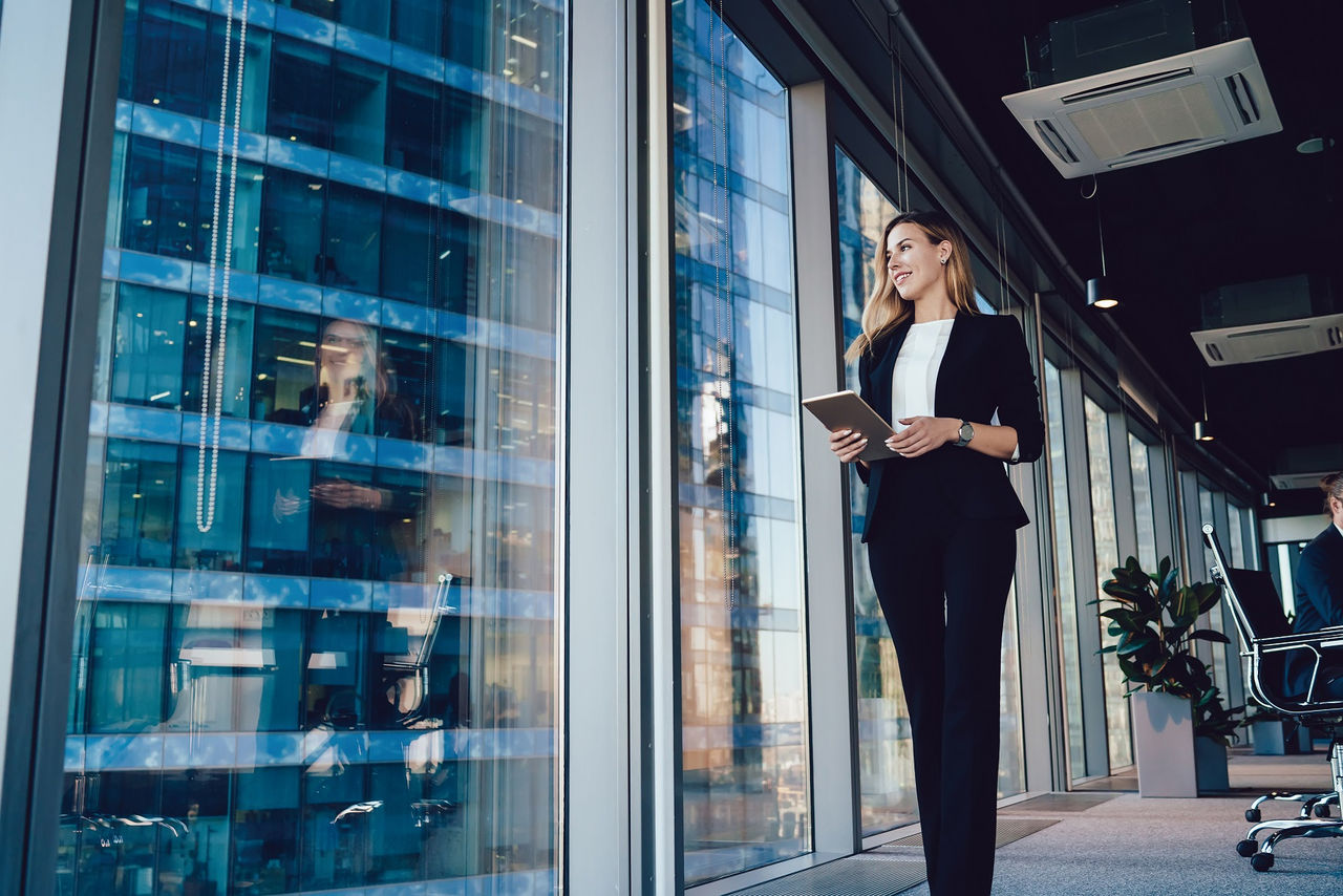 Caucasian female entrepreneur dressed in formal wear going near panoramic window in office, confident business woman with modern digital tablet looking away during working day in enterprise