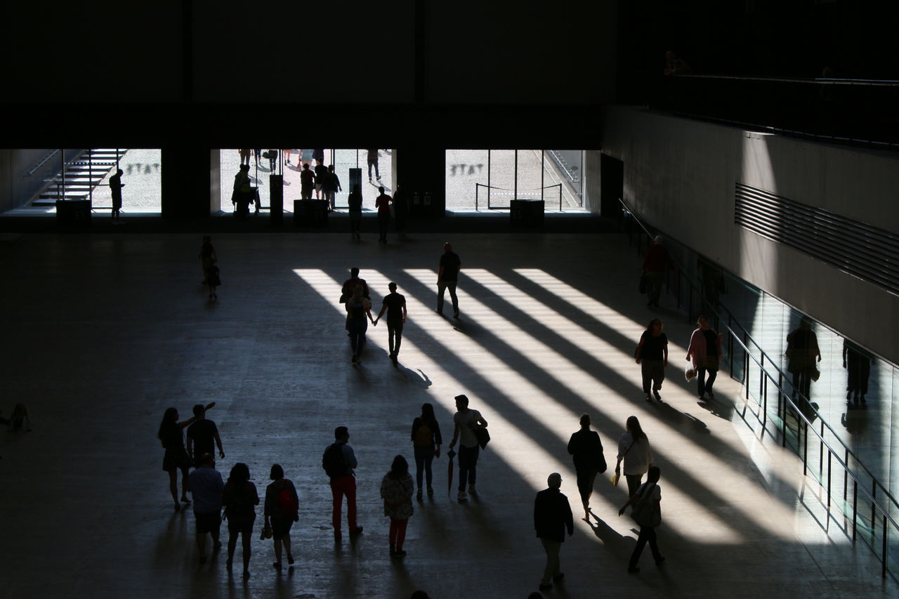 Interior of urban art gallery vast open space with people moving walking through, columns of light streaming white as blocks against dark shadow with figures silhouetted in Tate Modern entrance London, Interior of urban art gallery vast open space with people moving